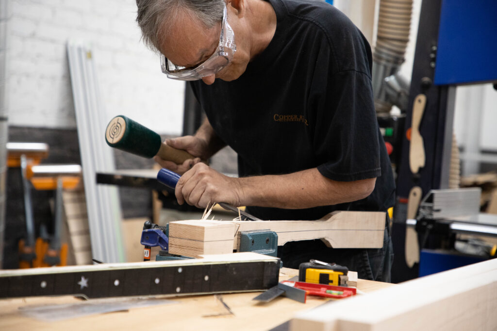 A man in a wood shop uses a rubber mallet and chisel to fabricate a guitar neck.