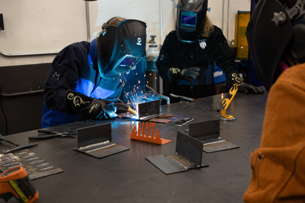 Two people wearing welding helmets and gloves work at a metal table, with one actively welding a steel joint as sparks fly, while small welded metal pieces sit arranged on the work surface in a makerspace workshop.